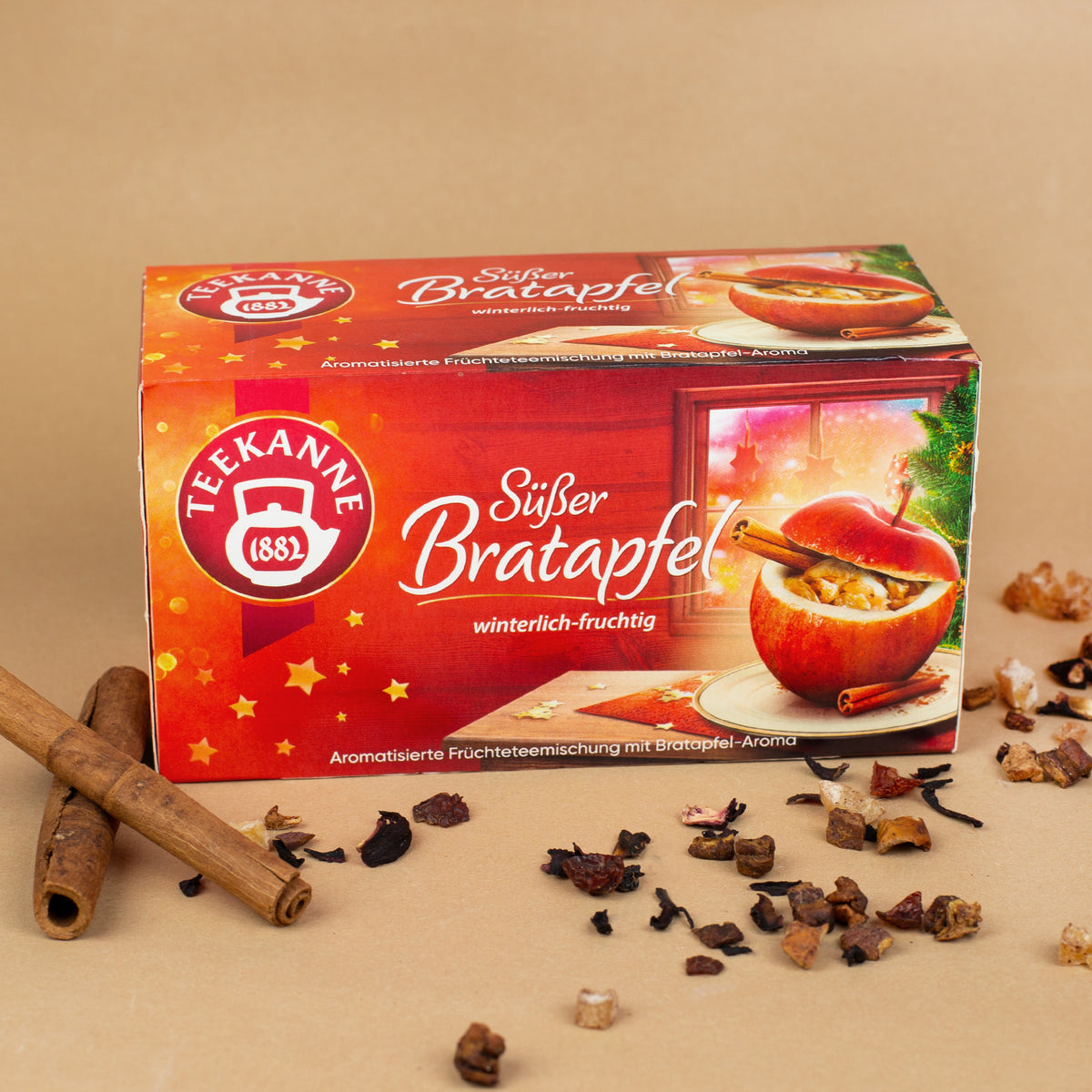 Schräg fotografierte Packung „Teekanne Süßer Bratapfel“ umgeben von losen Teestückchen, Zimtstangen und winterlicher Deko.