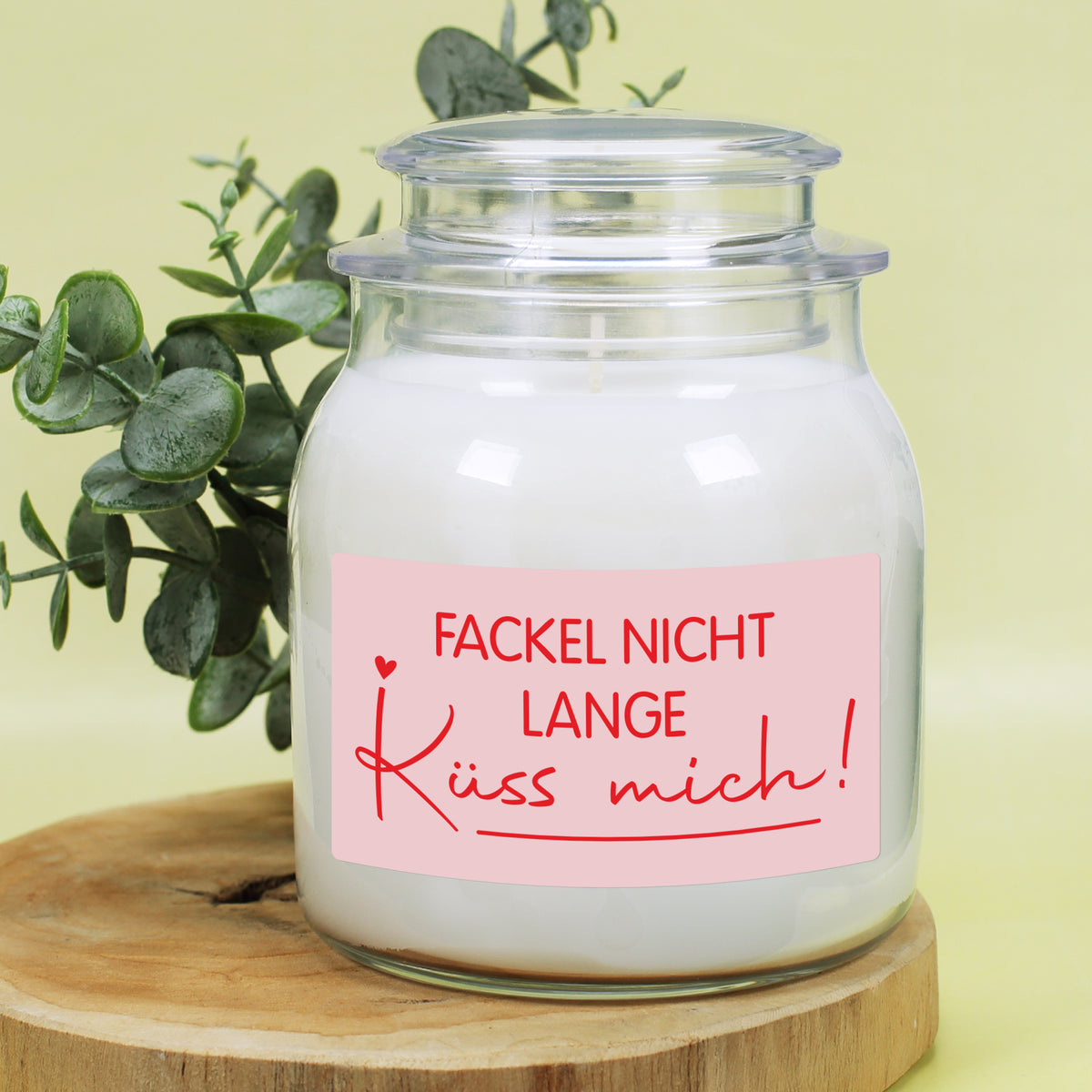 Weiße Duftkerze im Glas mit Deckel und romantischem Etikett „Fackel nicht lange – Küss mich!“ vor hellem Hintergrund