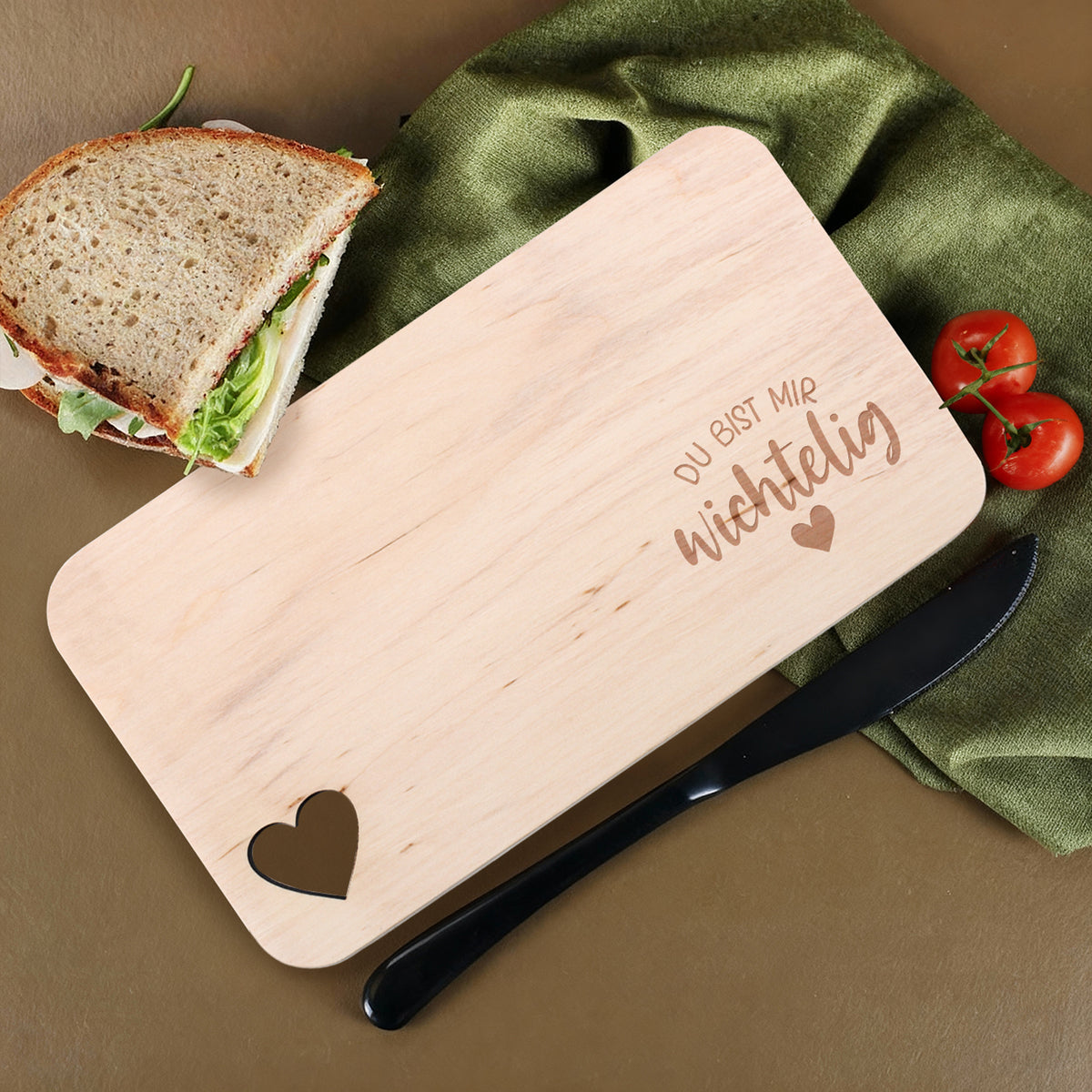 Holzbrettchen mit Gravur „Du bist mir wichtelig“ und Herzausschnitt, dekoriert mit Sandwich und Tomaten.