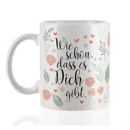 Weiße Keramiktasse mit floralem Dekor und Spruch „Wie schön, dass es Dich gibt“ auf heller Fläche stehend.