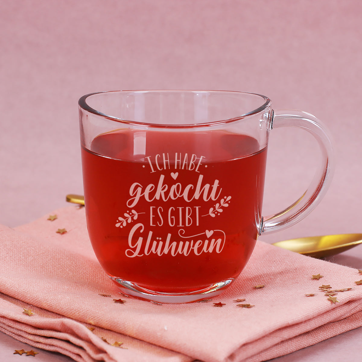 Leonardo Glastasse mit Gravur „Ich habe gekocht – es gibt Glühwein“ gefüllt mit rotem Glühwein auf rosa Stoffserviette mit goldenen Sternen