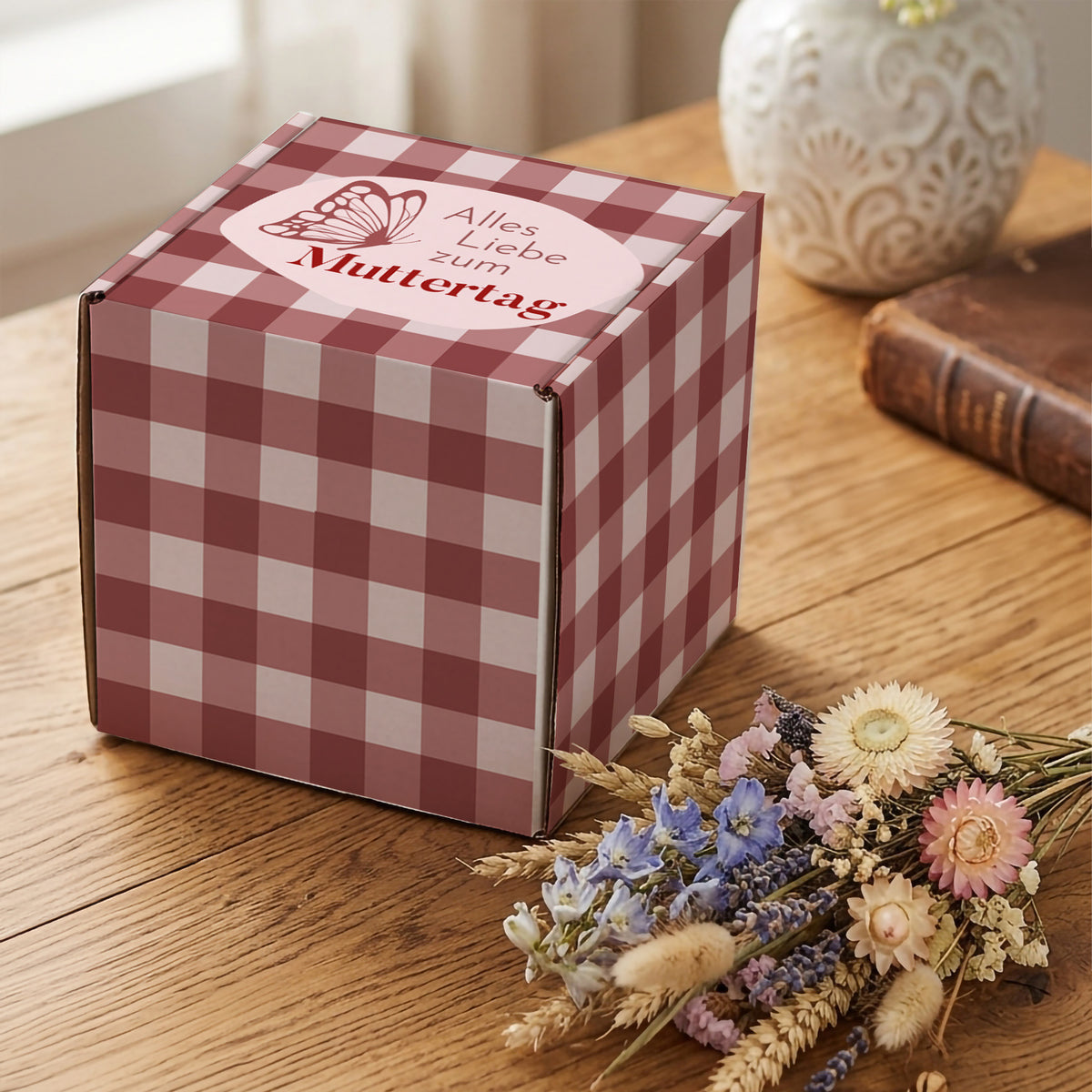 Muttertags Geschenkbox mit rosa Karomuster und Schmetterlingsmotiv auf Holztisch neben Trockenblumen