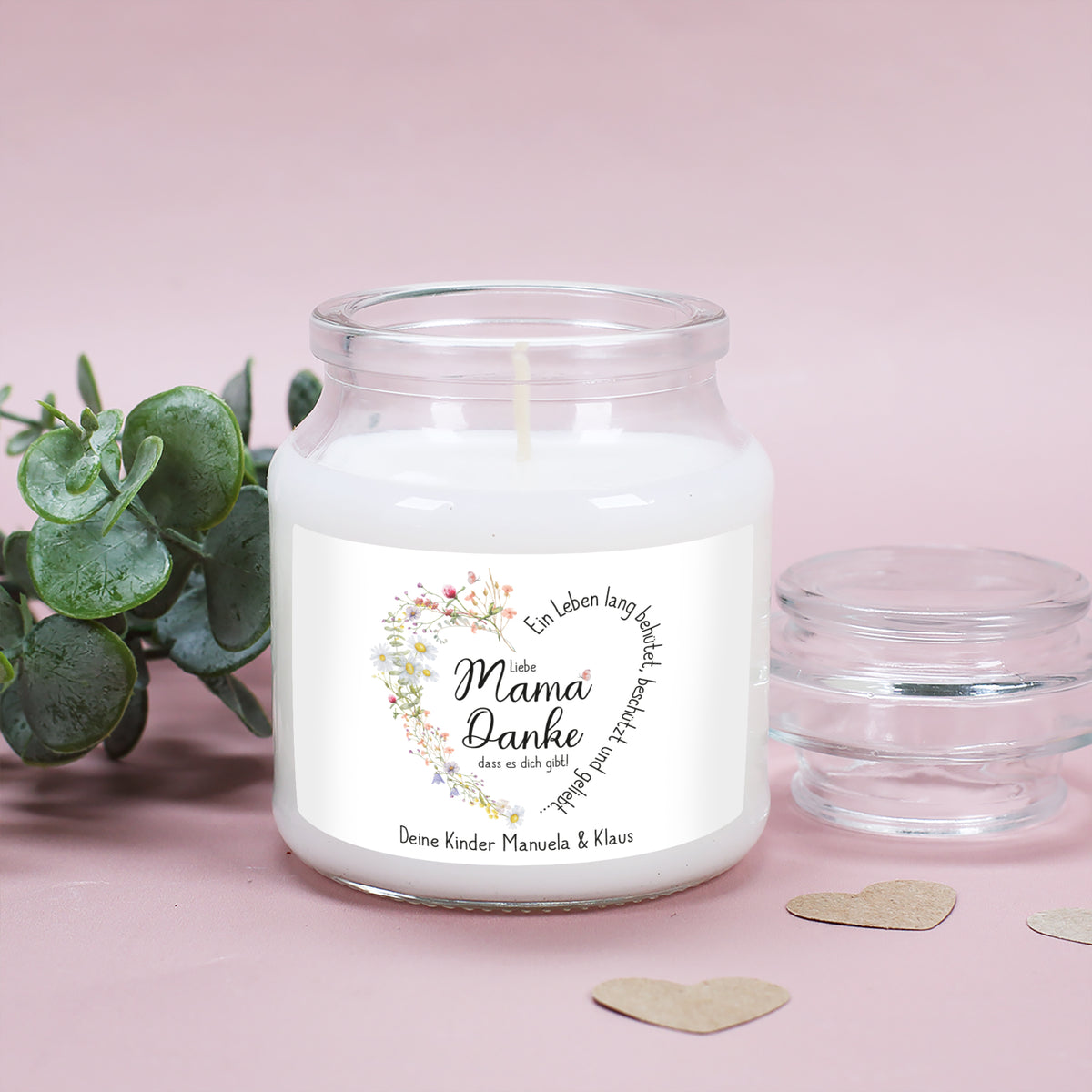 Duftkerze im stabilen, wiederverwendbaren Glas mit Deckel, platziert auf einer sanften rosa Unterlage mit grünen Eukalyptusblättern. Das Etikett zeigt ein florales Herz aus feinen Wiesenblumen mit dem Spruch „Ein Leben lang behütet, beschützt und geliebt…“. In der Mitte steht „Liebe Mama, Danke, dass es dich gibt!“. Die untere Textzeile ist personalisiert mit „Deine Kinder Manuela & Klaus“. Ein persönliches und stilvolles Geschenk für Mütter.