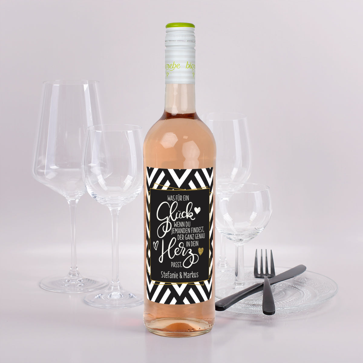 Roséweinflasche mit schwarz-weißem Etikett und goldener Schrift, Spruch „Was für ein Glück…“ und Namen Stefanie & Markus, vor Gläsern und Besteck dekoriert