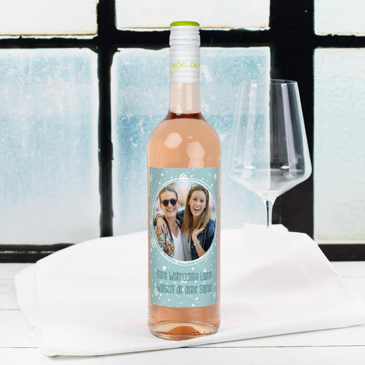Personalisierte Roséweinflasche mit Schneeflocken-Etikett, eigenem Foto und Weihnachtsgruß – Szene vor Fenster