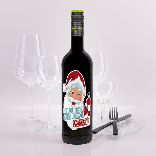 Weinflasche mit rotem Wein, Etikett Weihnachtsmann-Motiv „Weil du sooo artig warst! Konrad“, festlich dekoriert mit Gläsern.