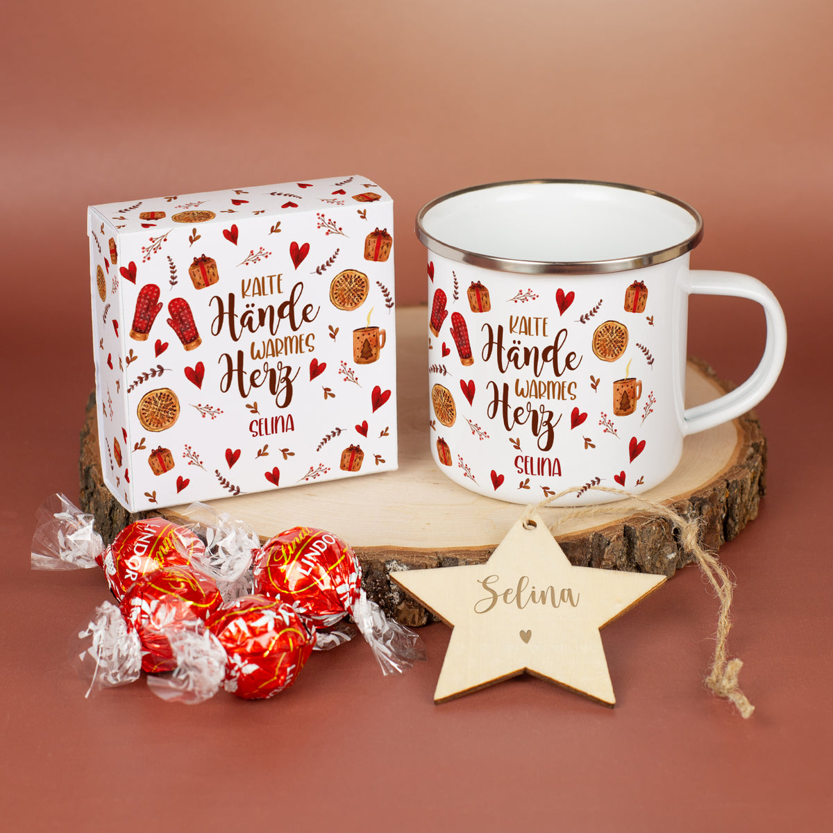 Nahaufnahme des Inneninhalts: Personalisierte Metalltasse mit Spruch „Kalte Hände warmes Herz“ und Name „Selina“. Daneben die dazugehörige Geschenkbox mit vier Lindorkugeln im gleichen Motivdesign sowie ein gravierter Holzstern mit Namensgravur „Selina“, arrangiert auf einer Holzscheibe.