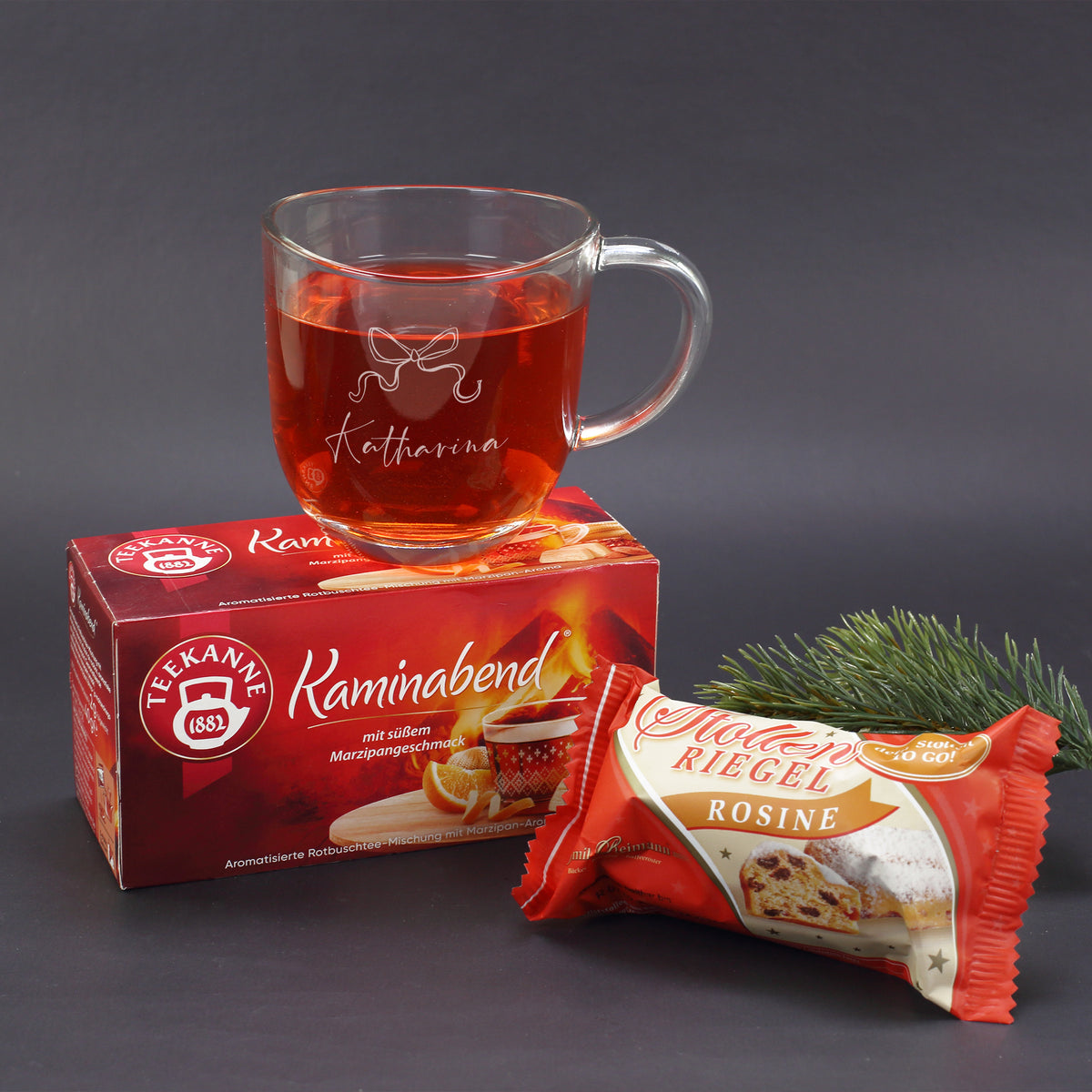 Geschenkset mit personalisierter Glastasse, Tee und Stollen vor dunklem Hintergrund und Tannenzweig