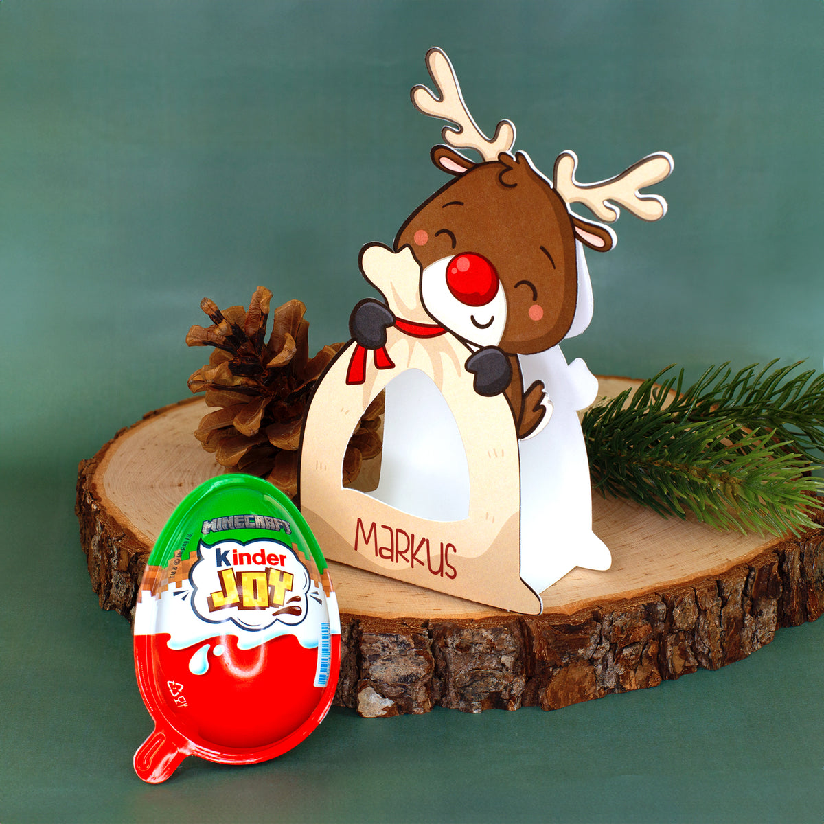 Personalisierte Rentier-Geschenkverpackung mit Name Markus neben Kinder Joy auf weihnachtlicher Holzdekoration