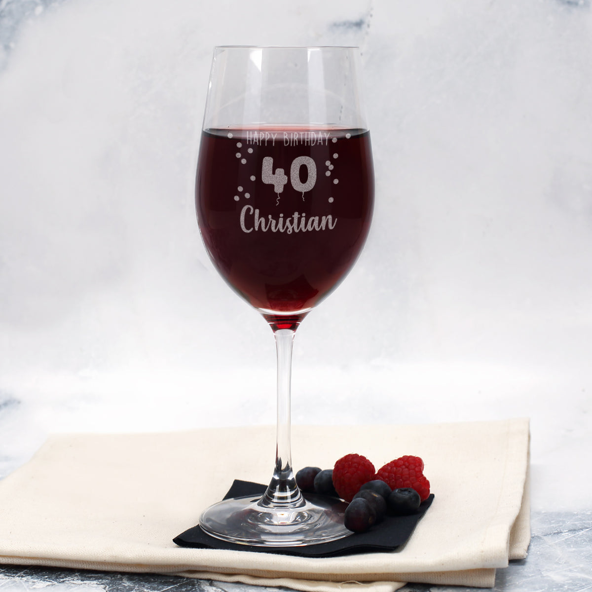 Graviertes Leonardo-Weinglas mit der Aufschrift „Happy Birthday 40 Christian“, gefüllt mit Rotwein, daneben Beeren und ein schwarzes Tuch