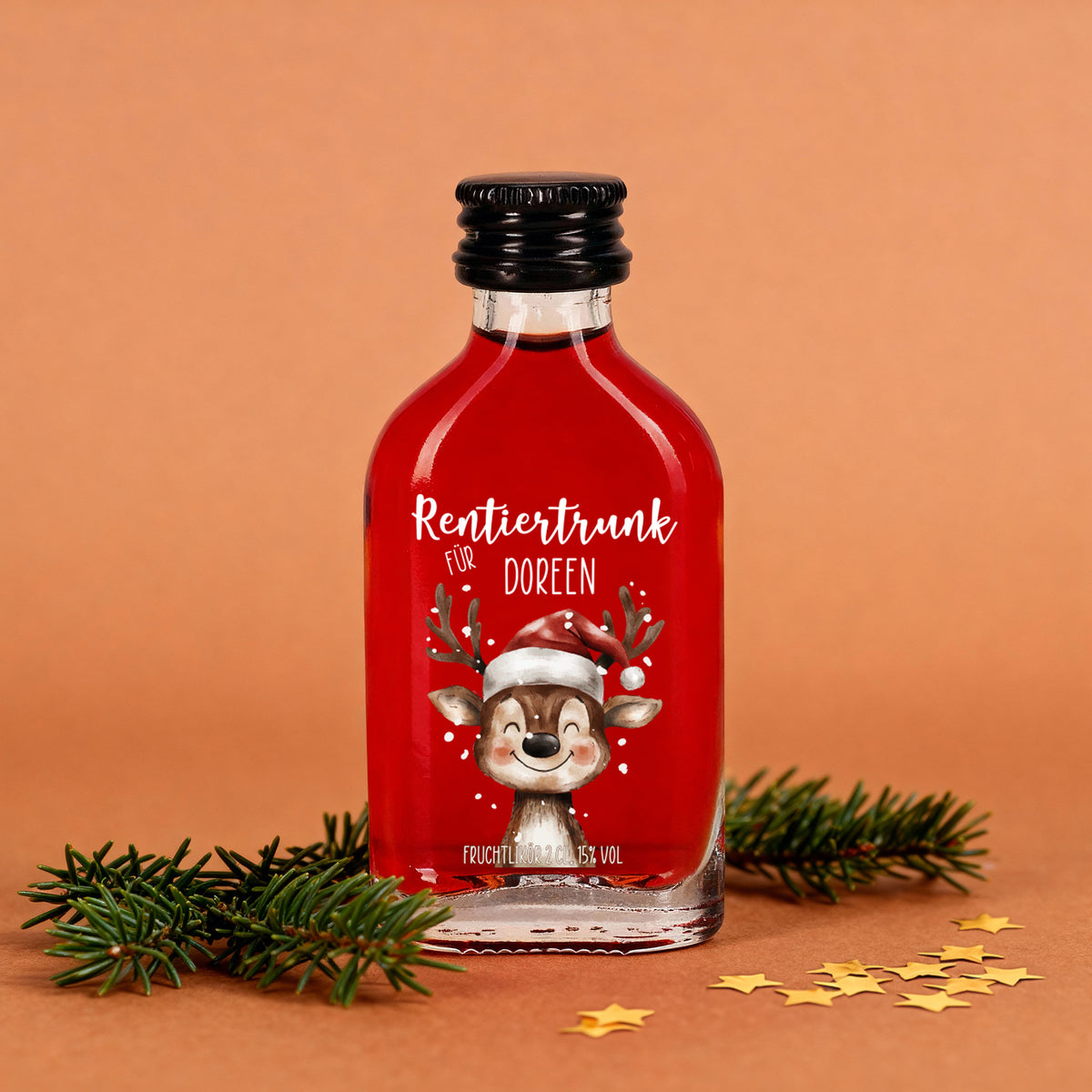 Kleine Glasflasche mit rotem Fruchtlikör, Aufdruck „Rentiertrunk für Doreen“ und fröhlichem Rentier mit Weihnachtsmütze, dekoriert mit Tannenzweigen und goldenen Sternen.