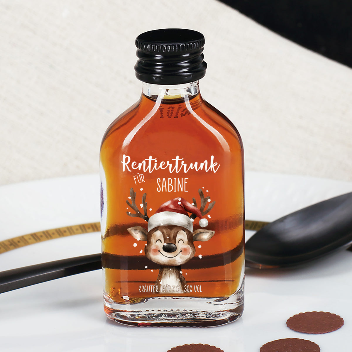 Kräuterlikör-Flasche „Rentiertrunk für Sabine“ auf weißem Teller mit schwarzem Löffel und braunen Papierpunkten, Fokus auf Etikett mit Rentier.