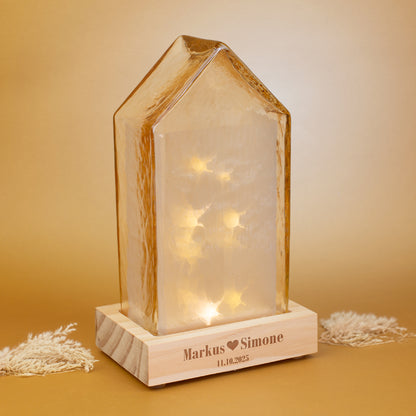 Amberfarbenes LED-Dekohaus von Leonardo auf Holzsockel mit personalisierter Gravur „Markus - Simone“, warm leuchtend im Hintergrund.