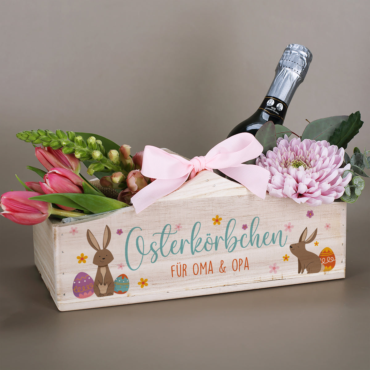 Personalisierter Pflanzkorb aus Holz zu Ostern mit Hasenmotiv und Aufdruck „Osterkörbchen“, dekoriert mit Blumen und Flasche