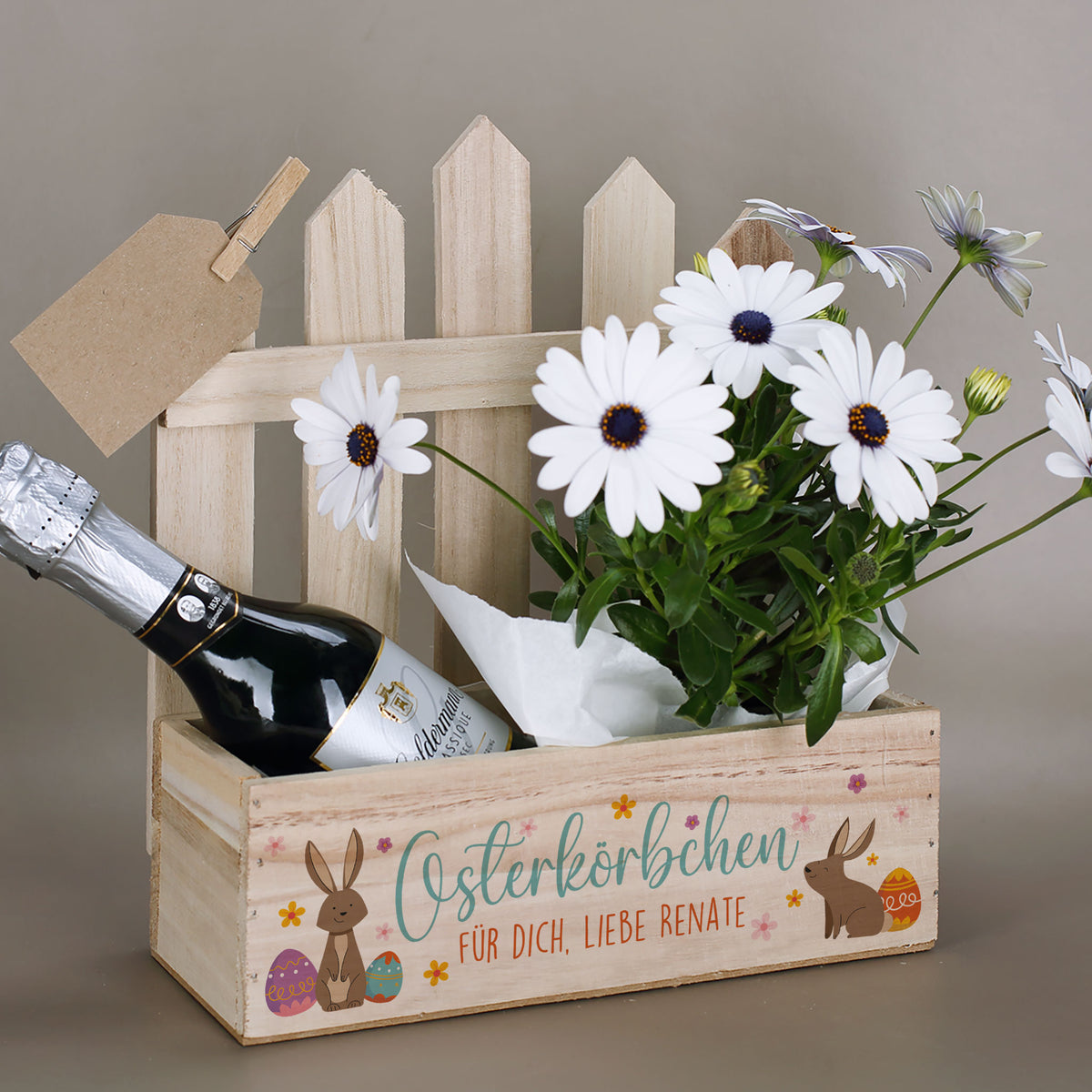 Holz-Pflanzkorb zu Ostern mit Zaunelement und Hasenmotiv, personalisiert mit Wunschtext, dekoriert mit Blumen und Flasche