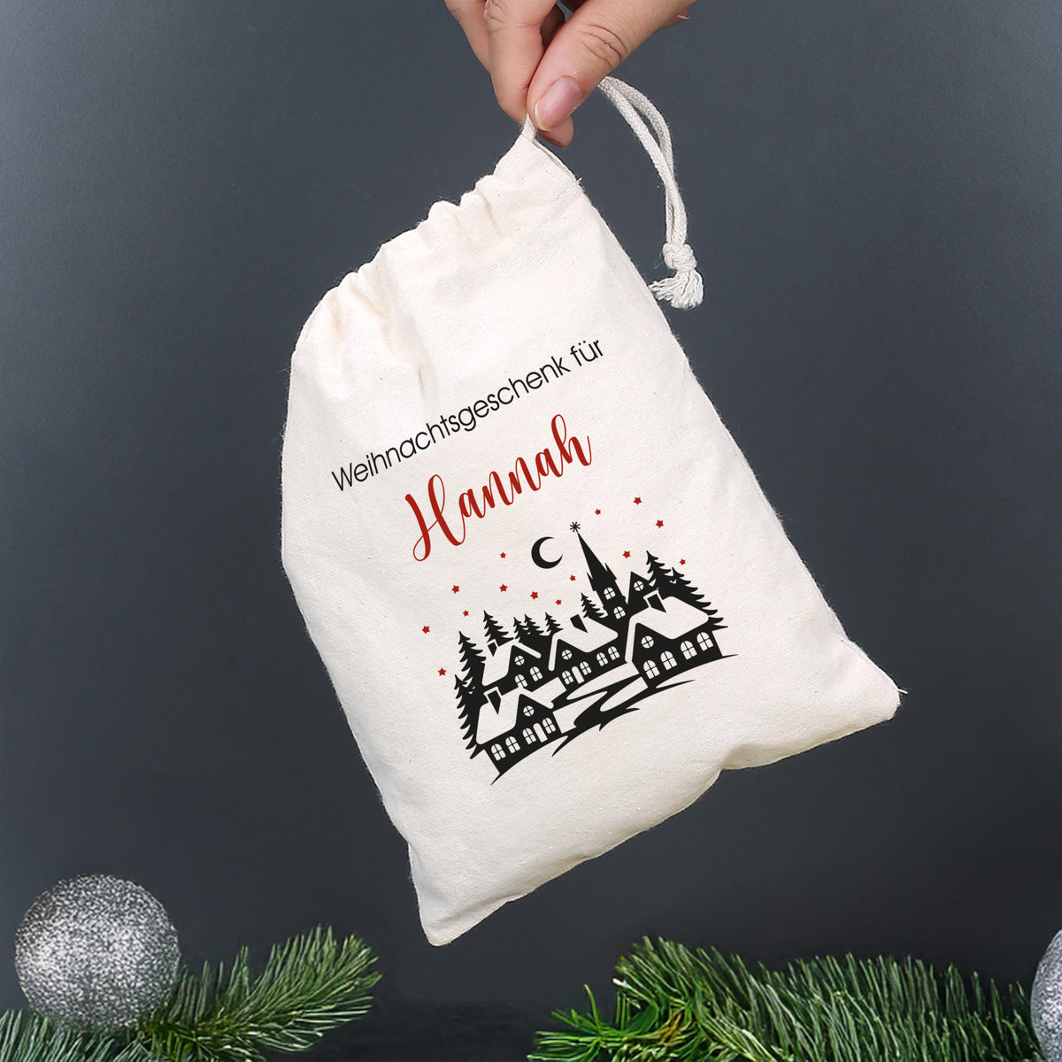 Hand hält einen kleinen Baumwollsack mit Wunschtext „Weihnachtsgeschenk für Hannah“, Wintermotiv und festlicher Hintergrund mit Kugeln.