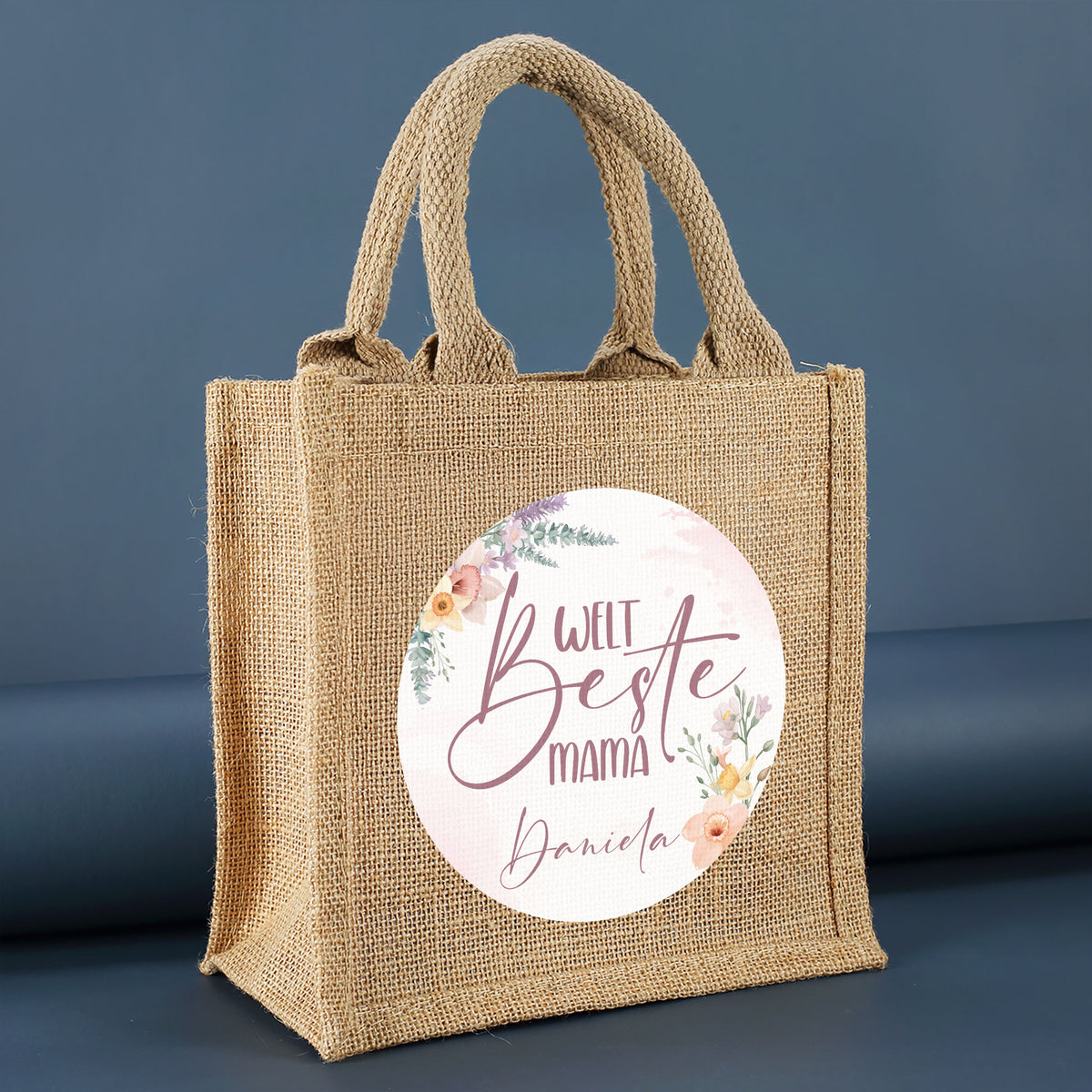 Jute-Geschenktasche „Weltbeste Mama“ mit floralem Motiv & personalisierbarem Namen seitlich fotografiert auf dunkelblauem Hintergrund