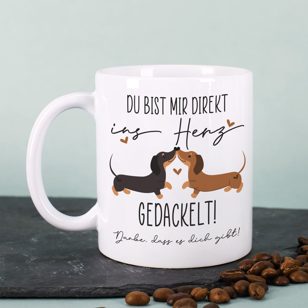 Keramiktasse mit zwei Dackeln und Herzsymbol, Spruch „Du bist mir direkt ins Herz gedackelt!“ auf dunkler Platte mit Kaffeebohnen