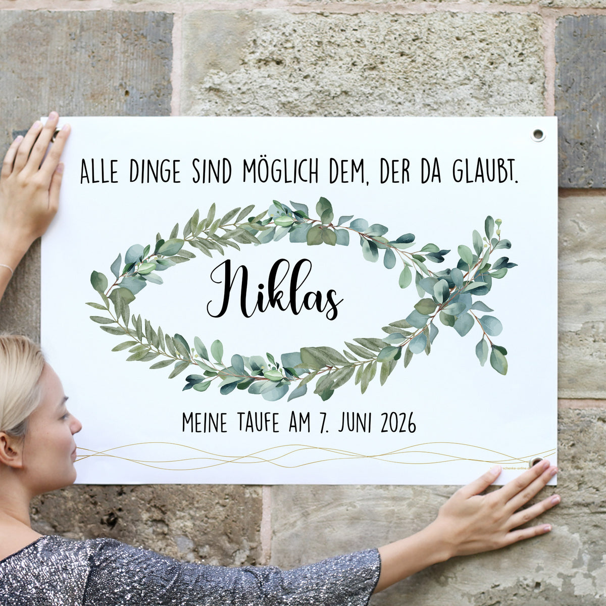 Outdoor-Banner mit Eukalyptus-Fischmotiv und dem Text „Alle Dinge sind möglich dem, der da glaubt.“ sowie Name „Niklas“ und Datum „Meine Taufe am 7. Juni 2026“. Eine Frau befestigt das Banner an einer Steinwand.