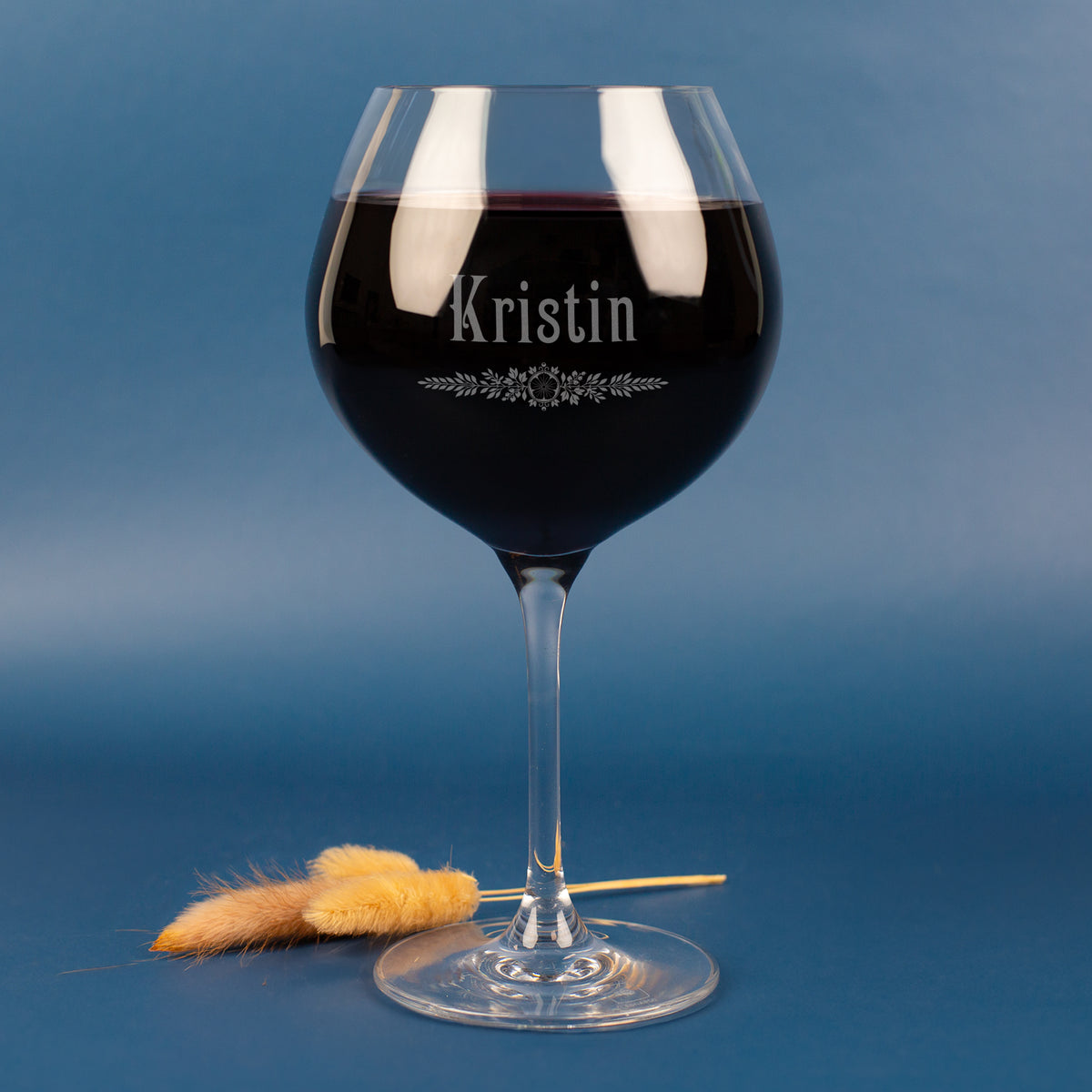 Personalisiertes Leonardo Weinglas mit Gravur des Namens Kristin und floralem Ornament – edles Burgunderglas mit Rotwein, vor blauem Hintergrund mit zarten Trockenhalmen.