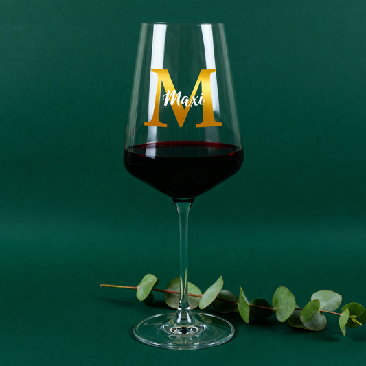 Personalisiertes Leonardo Weinglas mit goldener Initiale M und Name Maxi, gefüllt mit Rotwein vor grünem Hintergrund