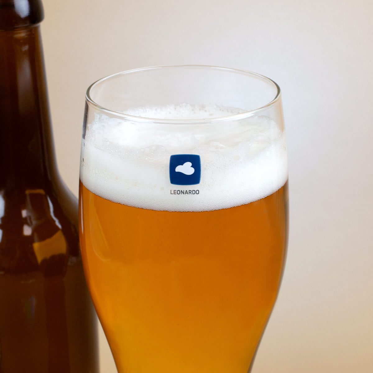 Detailaufnahme des Leonardo-Logos auf einem gefüllten Weizenglas mit Schaumkrone, im Hintergrund eine braune Bierflasche.