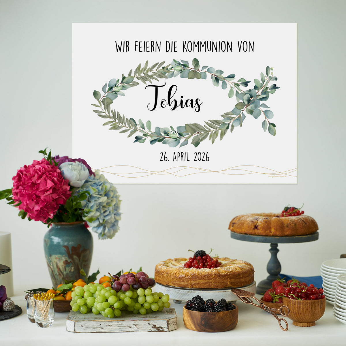 Poster-Banner mit christlichem Eukalyptus-Fischmotiv, Name „Tobias“ und Datum „26. April 2026“. Das Banner hängt über einem festlich gedeckten Tisch mit Kuchen, Obst und Blumen.