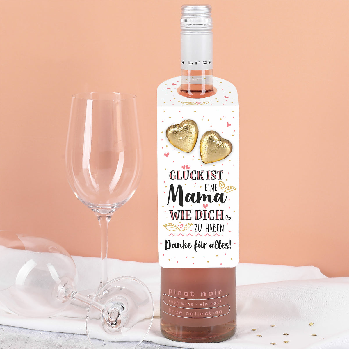 Roséweinflasche mit Muttertags-Flaschenanhänger, Text und zwei goldenen Lindt-Schokoherzen, daneben Weingläser