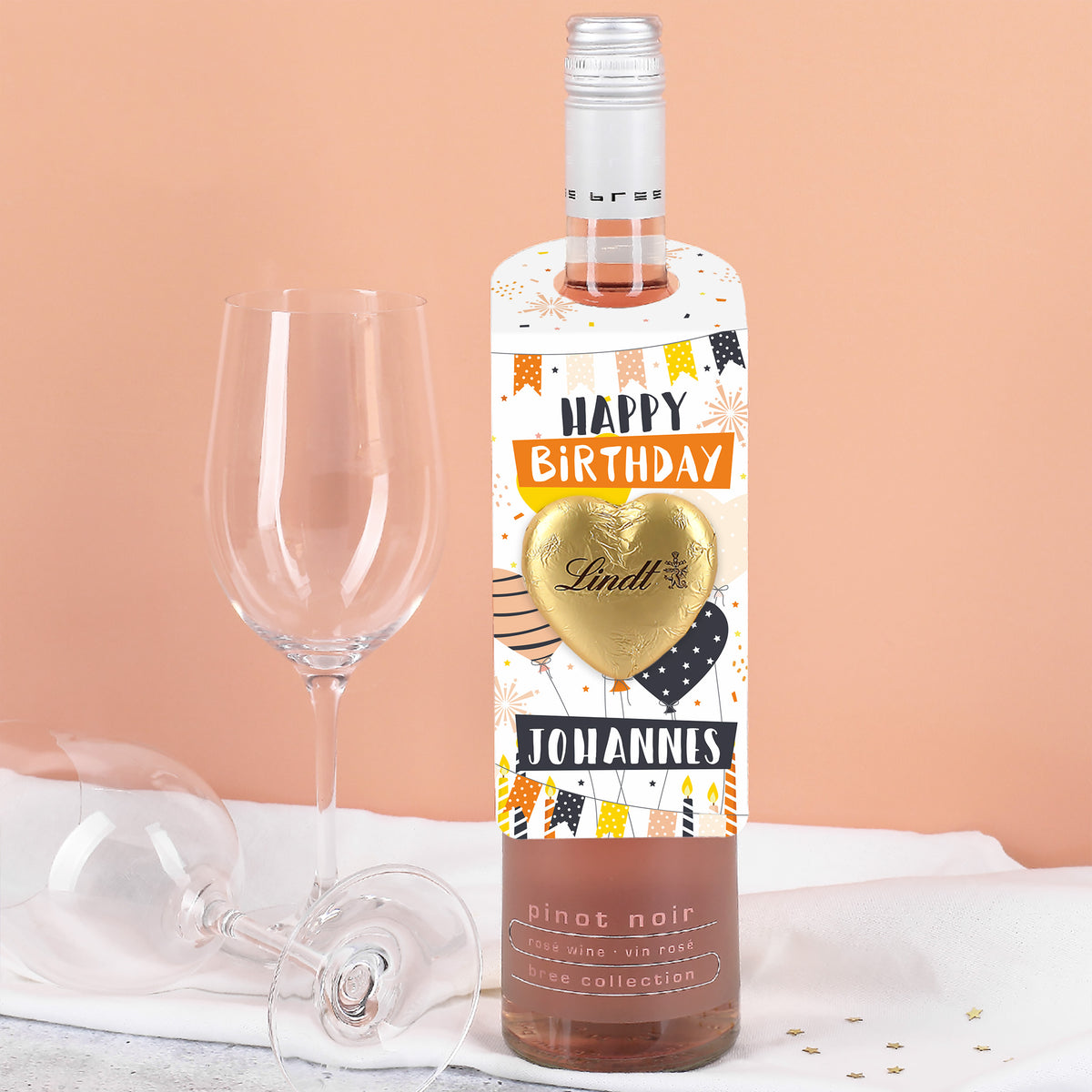 Flaschenanhänger mit „Happy Birthday“-Design in Weiß, Orange und Schwarz, goldfarbenes Lindt Schokoladenherz und personalisierter Name – an Flasche befestigt.