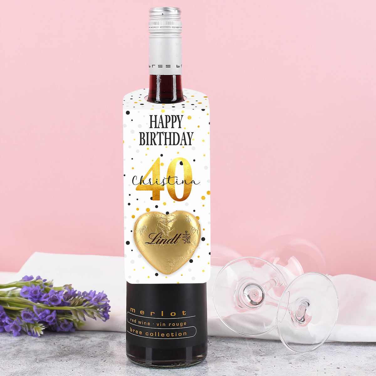 Großer Flaschenanhänger zum Geburtstag mit Konfetti-Motiv, personalisiertem Namen und Alter sowie goldenem Lindt-Herz – auf einer Flasche platziert.