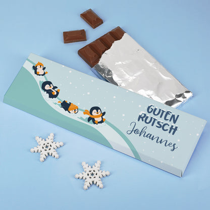 Schokoladentafel mit Pinguin-Motiv und Aufdruck „Guten Rutsch Johannes“ auf hellblauem Hintergrund, dekoriert mit Schneeflocken – Geschenkidee zum Jahreswechsel.