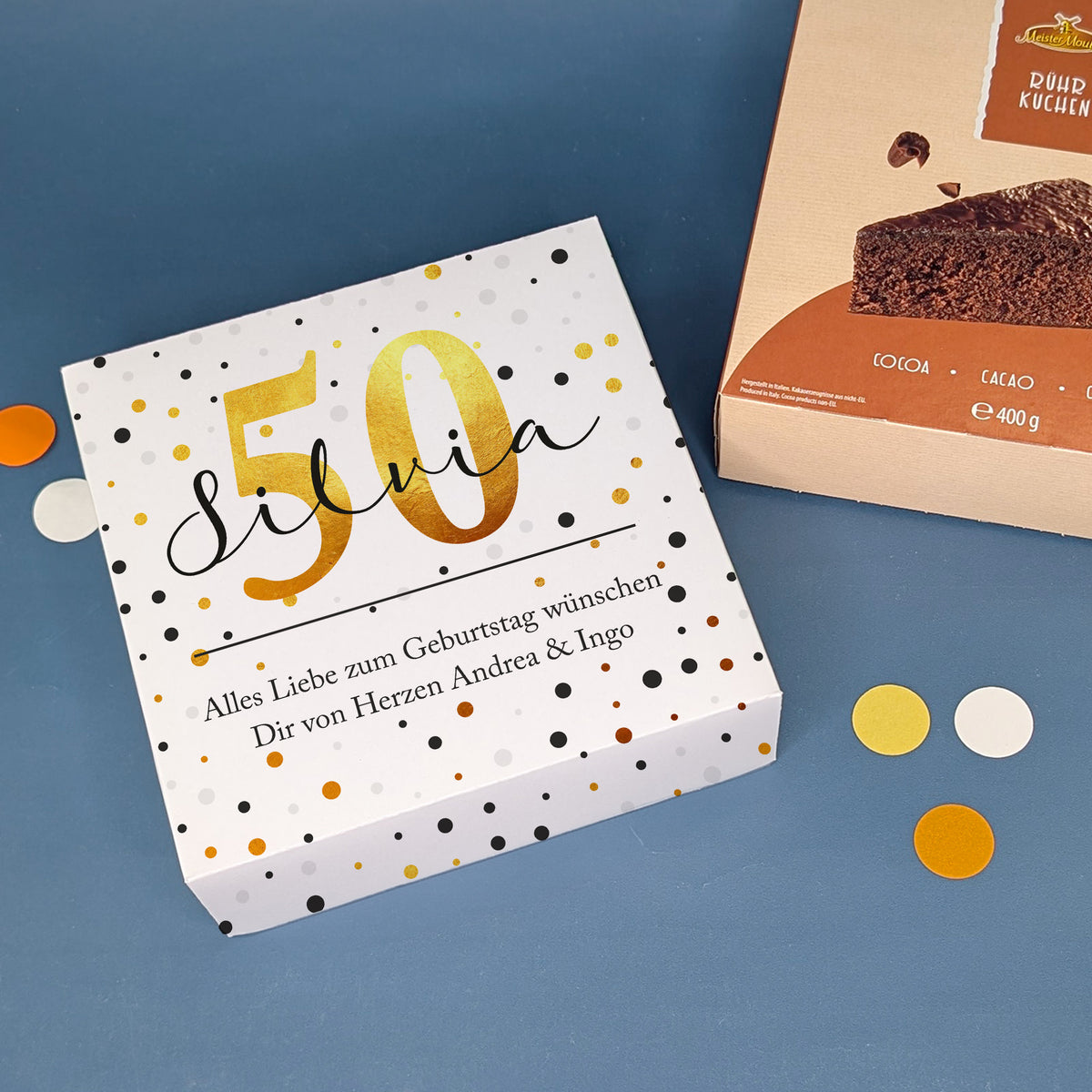 Geburtstagskuchen mit Zahl 50 und Name Silvia in Geschenkbox mit Spruch, daneben Schokokuchenpackung