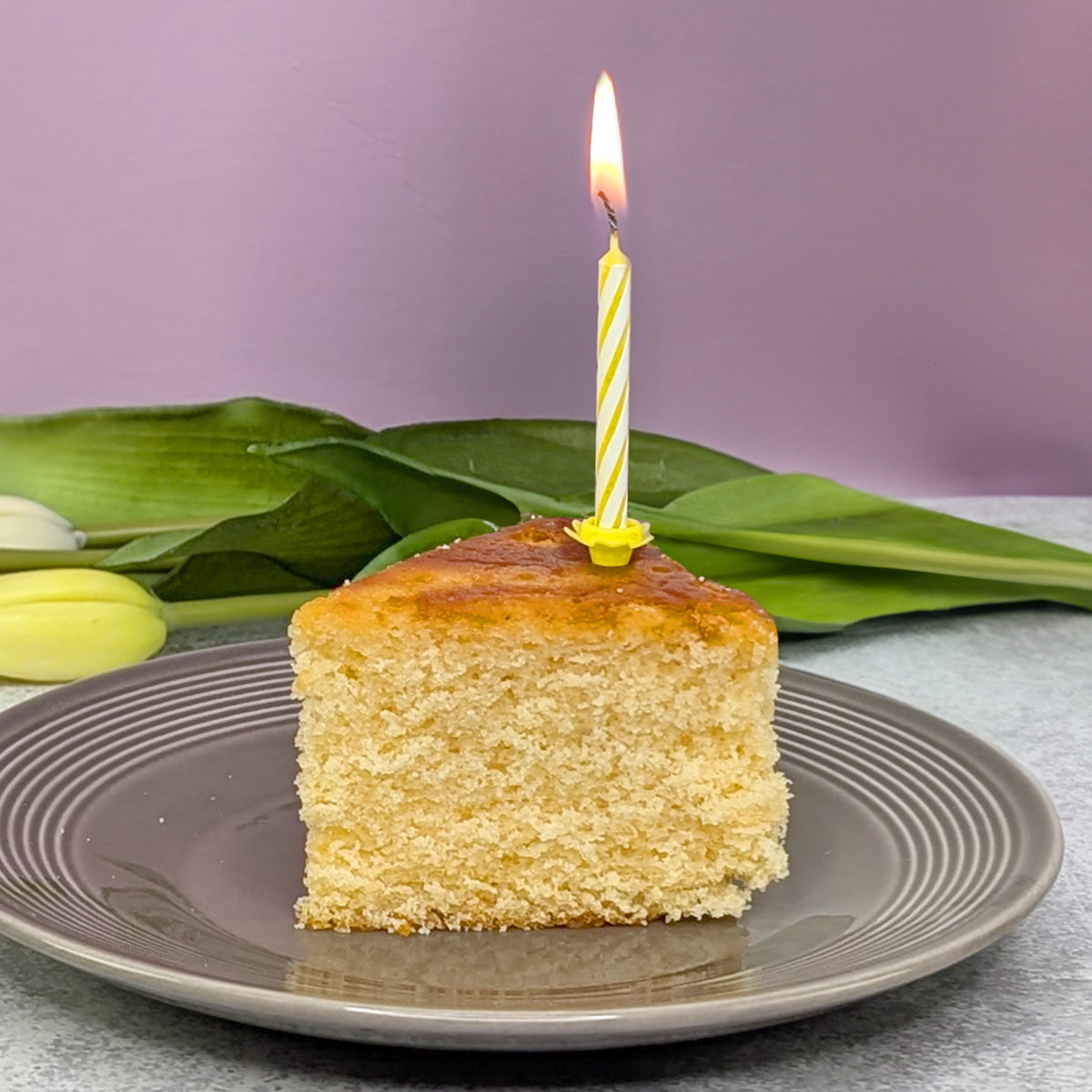 Zitronenkuchenstück mit brennender Kerze, im Hintergrund Tulpenblätter