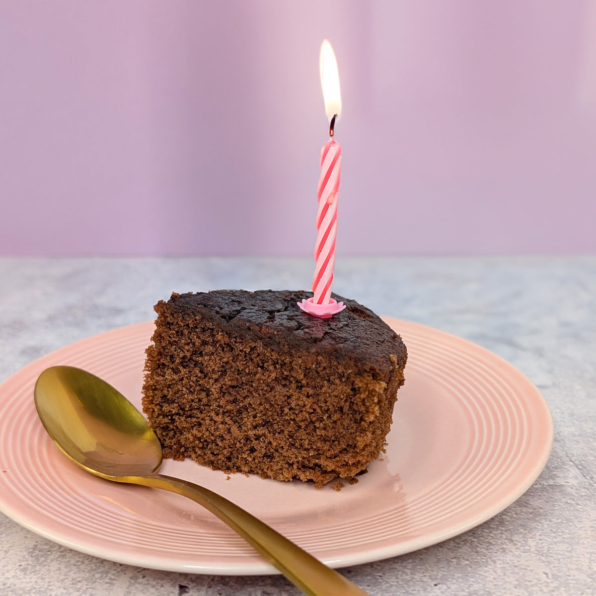 Stück Schokokuchen mit Geburtstagskerze auf rosa Teller, daneben goldener Löffel