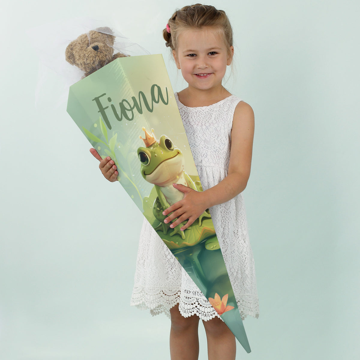 Mädchen in weißem Kleid lächelt mit einer großen Zuckertüte mit dem Namen "Fiona" und Frosch-Motiv