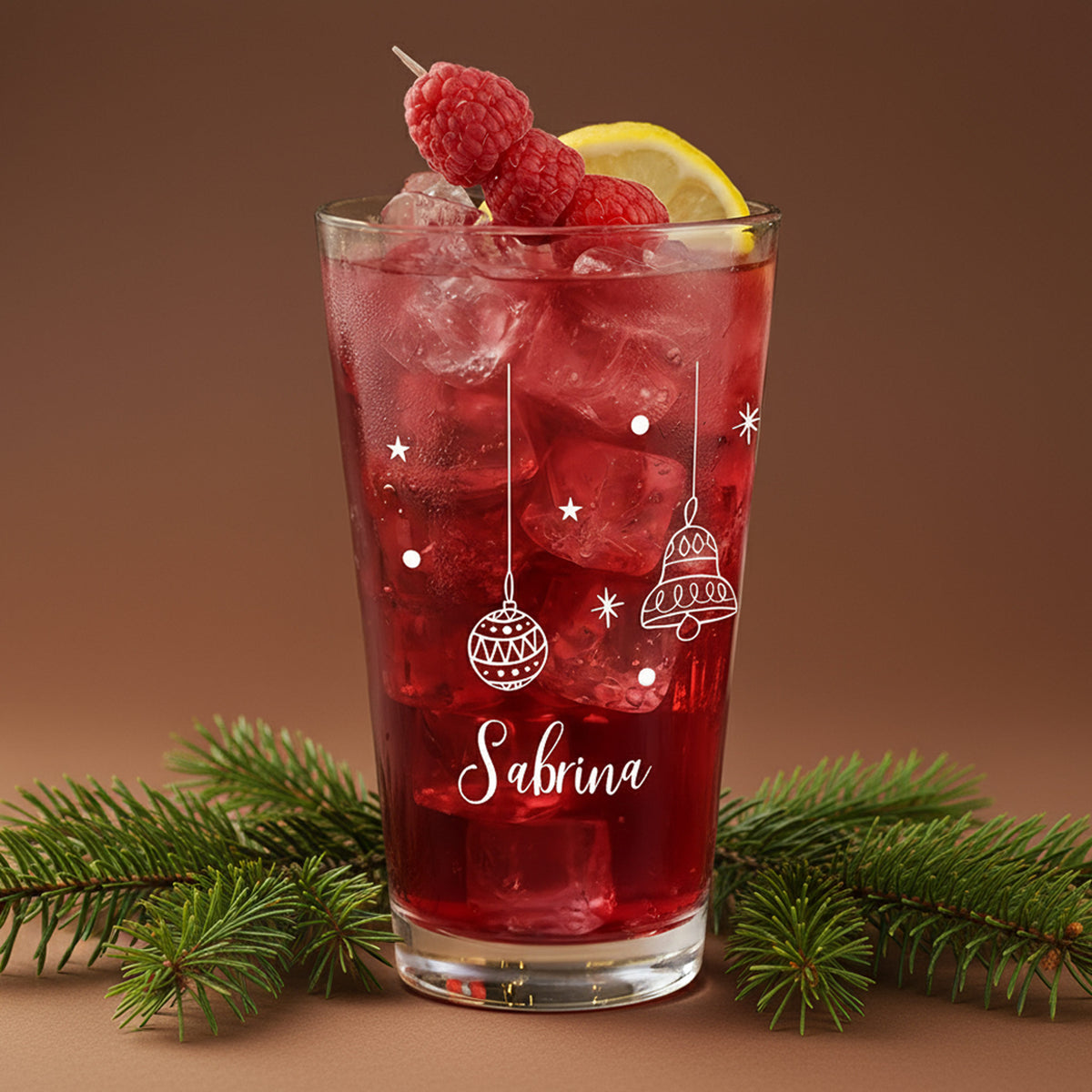 Festliches Trinkglas mit weißen Weihnachtsmotiven – Kugeln, Glocken und Sternen – befüllt mit rotem Getränk und Eis, mit weihnachtlicher Deko.