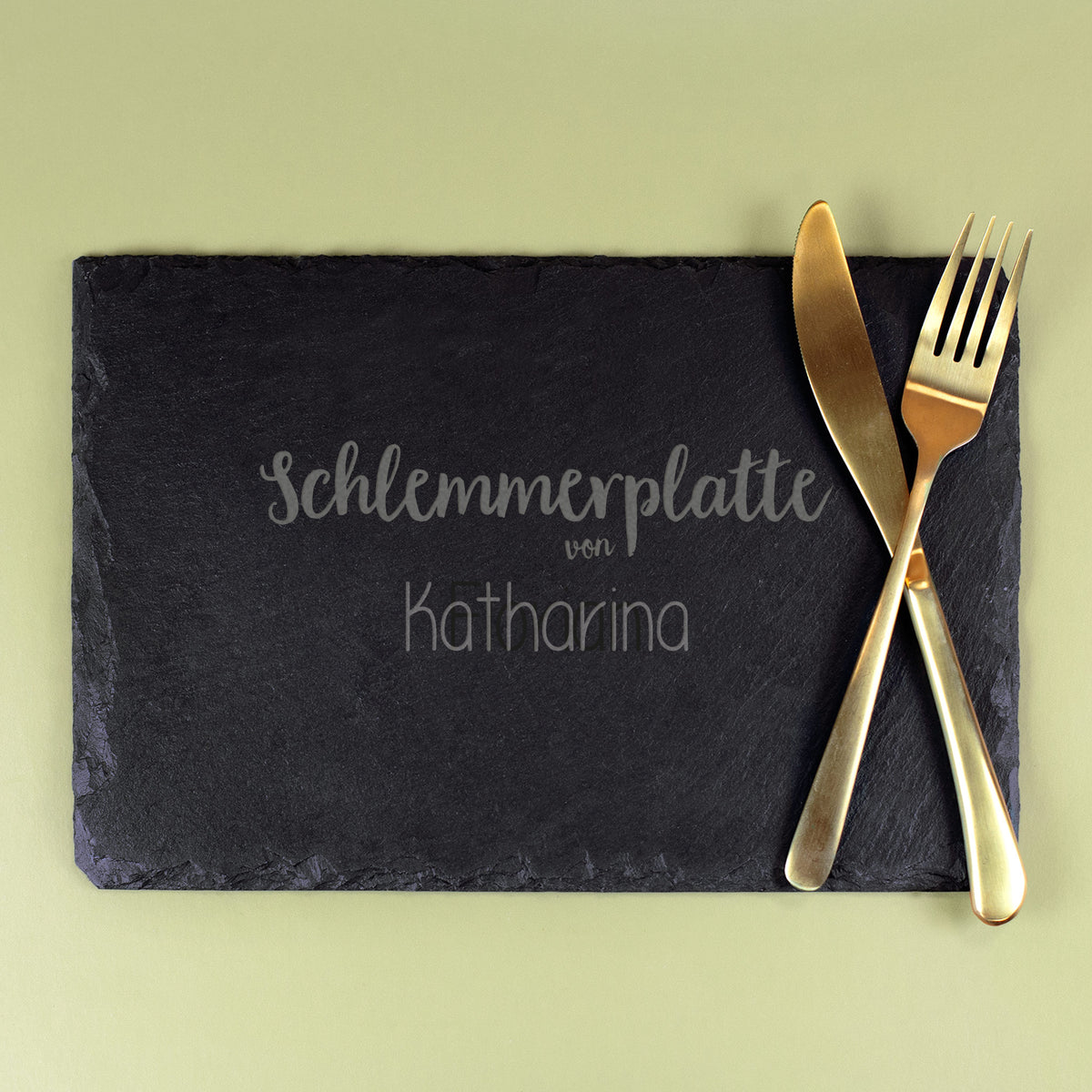 Schwarze Schieferplatte mit Gravur „Schlemmerplatte von Katharina“, daneben goldfarbenes Besteck (Messer und Gabel) auf grünem Untergrund.