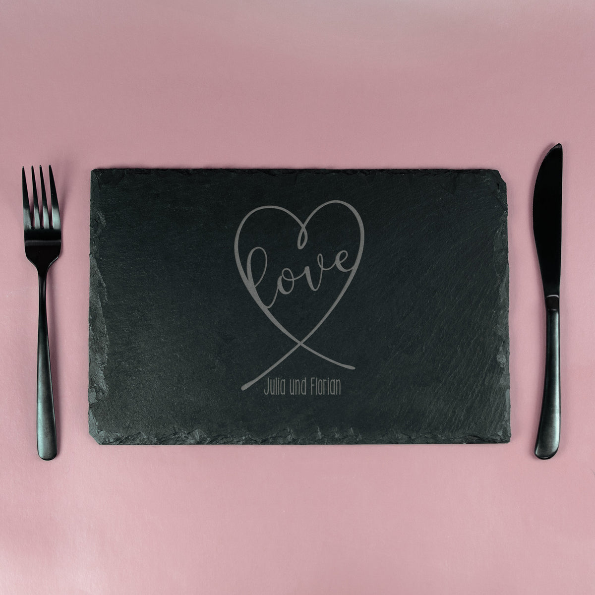 Schieferplatte mit graviertem Herz-Design und „Love“-Schriftzug, eingedeckt mit Besteck auf rosa Hintergrund
