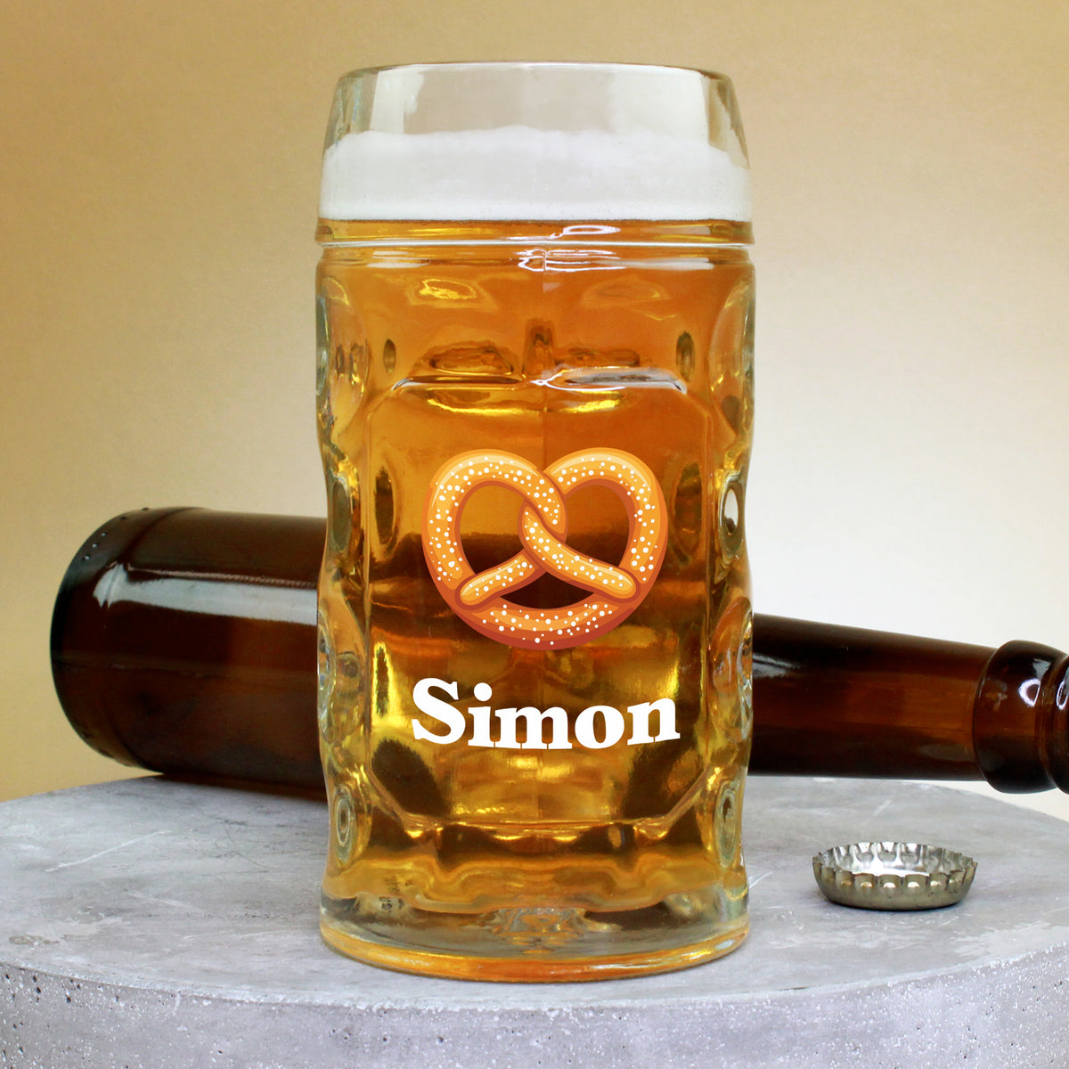 Bierglas mit Brezel-Motiv und Name Simon, aufrecht vor liegender Bierflasche und Kronkorken