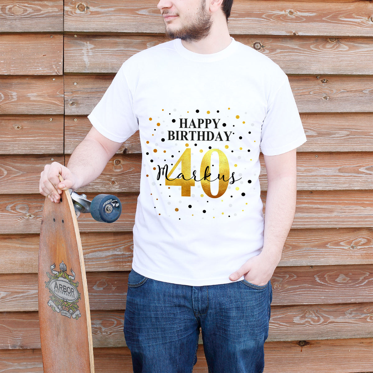 T-Shirt mit Geburtstags-Motiv & Schriftzug "Happy Birthday" personalisiert mit Alter & Name getragen von einem Model mit einem Longboard in der Hand vor einer Bretterwand