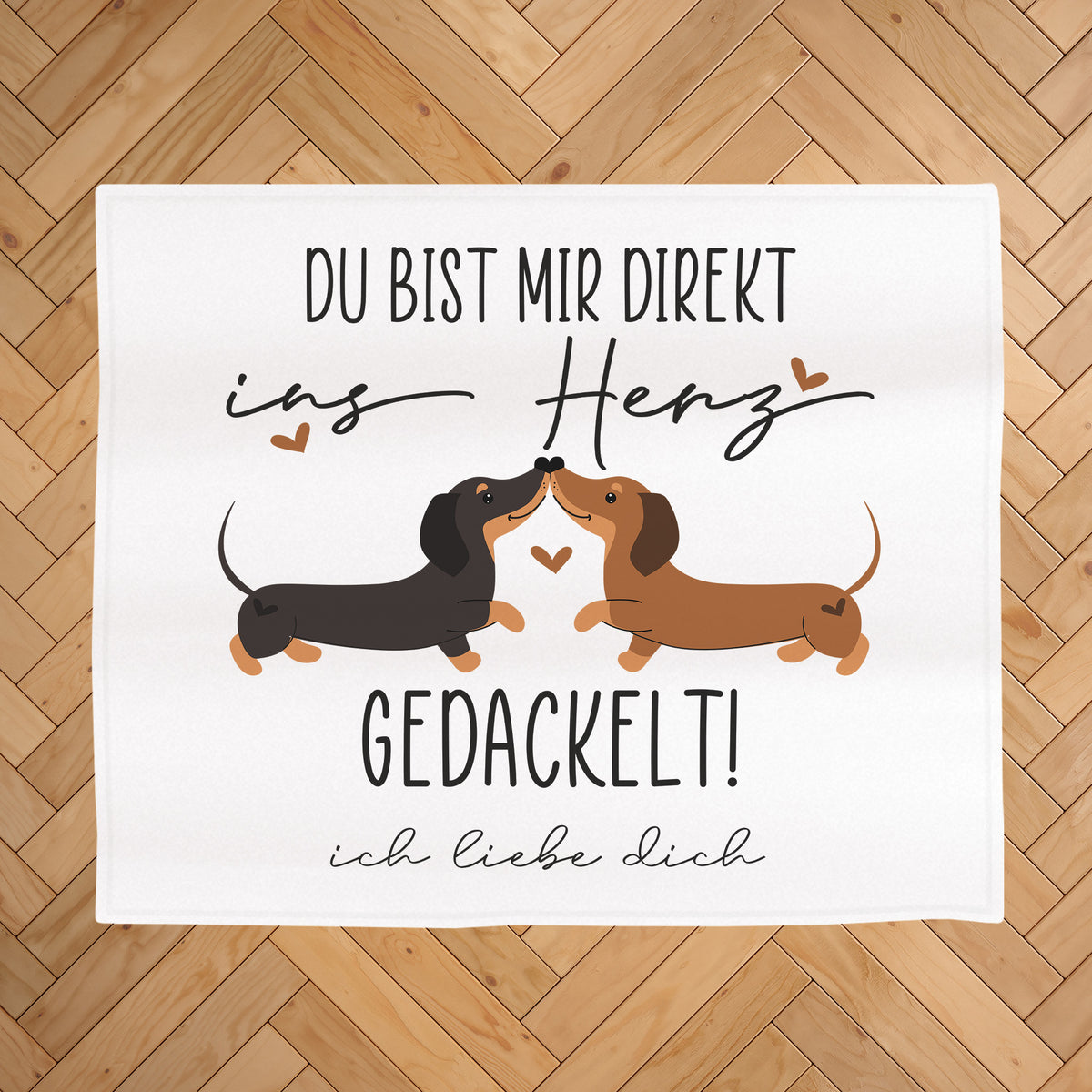 Kuscheldecke mit Dackel-Motiv auf hellem Holz-Fischgrätboden, Aufdruck „Du bist mir direkt ins Herz gedackelt!“ und Wunschtext „ich liebe dich“ unter dem Motiv