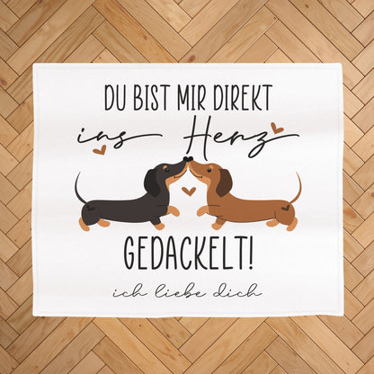 Kuscheldecke mit Dackel-Motiv auf hellem Holz-Fischgrätboden, Aufdruck „Du bist mir direkt ins Herz gedackelt!“ und Wunschtext „ich liebe dich“ unter dem Motiv