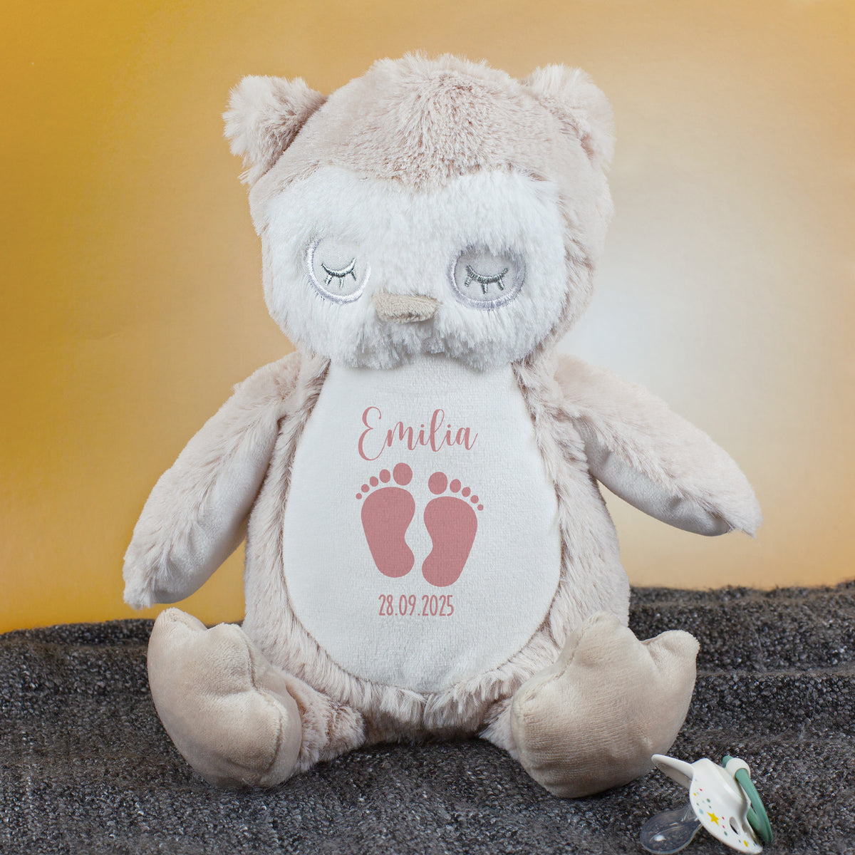 Eine sitzende Kuscheltier-Eule mit rosa Babyfüßchen-Motiv und der Personalisierung „Emilia – 28.09.2025“ auf ihrem Bauch. Das Plüschtier befindet sich auf einer weichen, dunklen Unterlage, daneben liegt ein Schnuller.