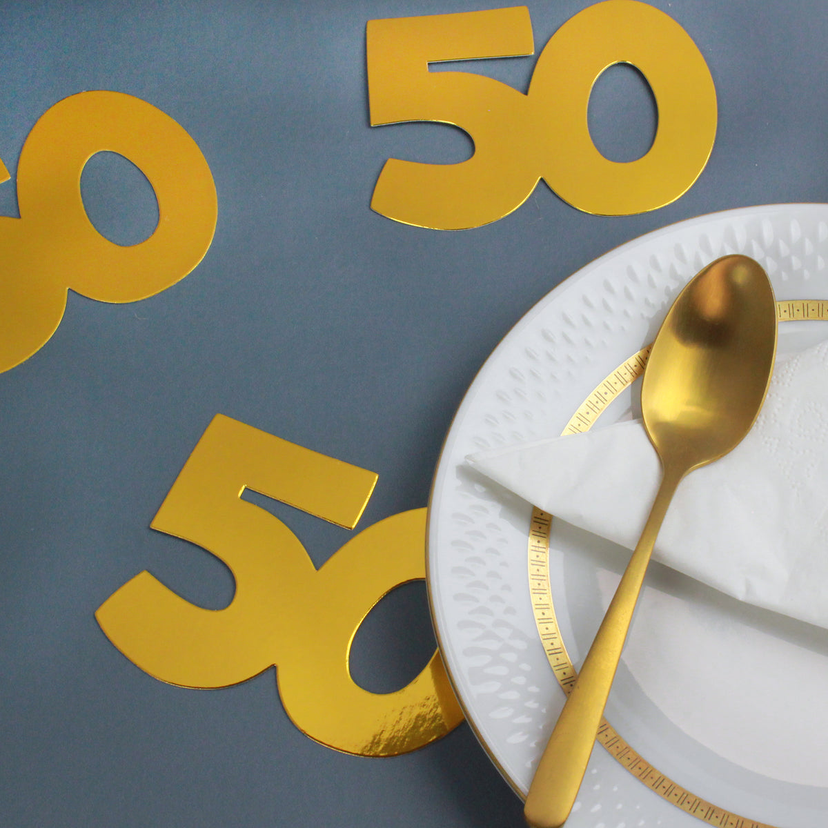 Goldene Jumbo-Konfetti Zahl 50 als Streudeko auf grauem Hintergrund neben Teller